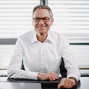 Professional portrait of a person seated at a desk, wearing a white button-down shirt and glasses, with hands folded on the tabletop in front of window blinds.