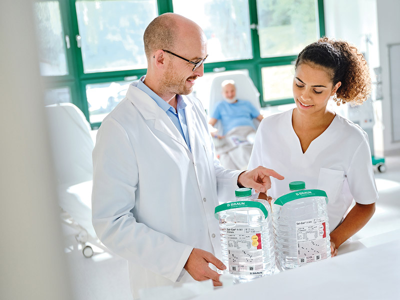 Two healthcare professionals, a nephrologist and a dialysis nurse, in a clinical setting review dialysis concentrate canisters with green caps, placed on a white surface. One person points at the canisters while both examine the labels. A patient is visible blurred in the background near large windows with natural light.