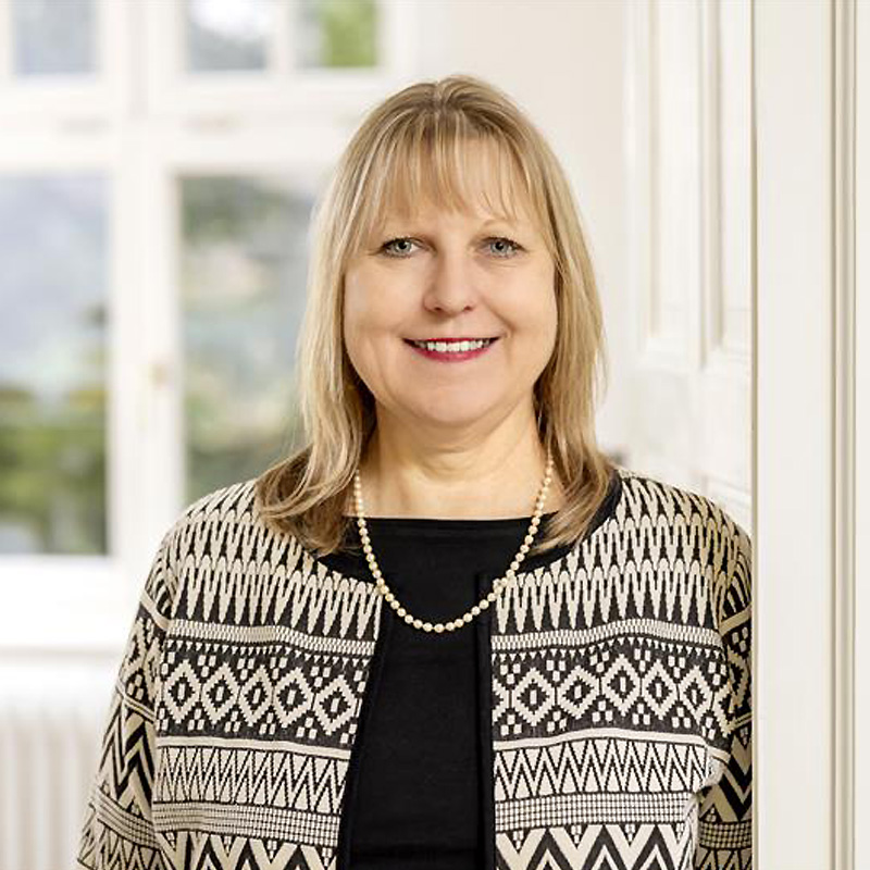 Jitka Pancířová, Senior Quality Manager Health Care Services at B. Braun Avitum, wearing a patterned cardigan over a black top with a pearl necklace, standing indoors near a bright window.