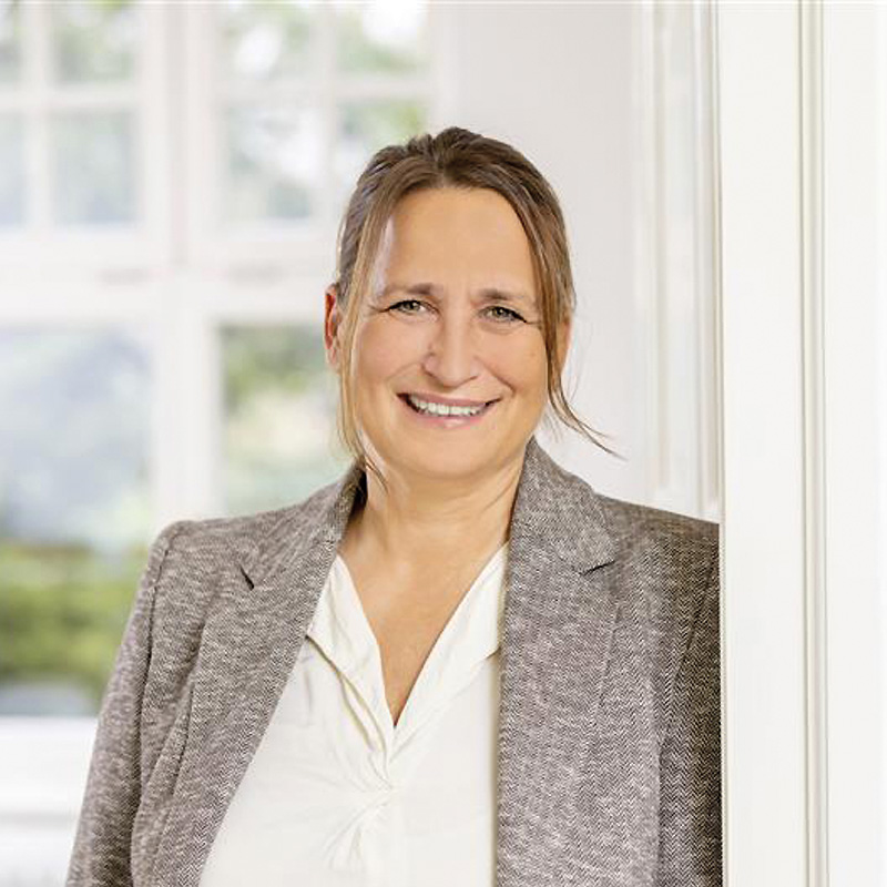 Simone Klein, Vice President Health Care Services at B. Braun Avitum, wearing a light blouse and a grey blazer, standing indoors near a bright window.