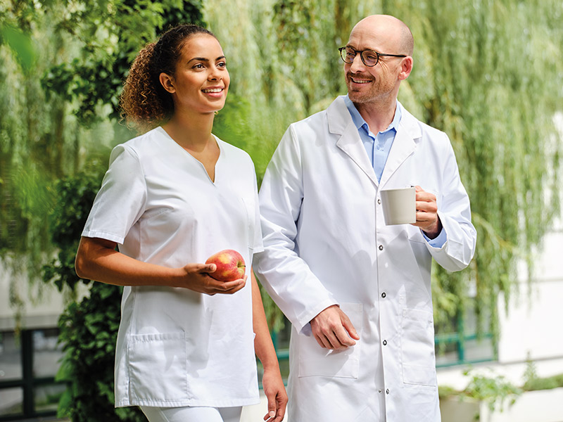 Two healthcare professionals, a nephrologist and a dialysis nurse, walk outdoors in a green, park-like setting. One person wears a white medical uniform and holds an apple, while the other wears a white lab coat and carries a mug. Trees and natural greenery form the background, creating a calm, fresh environment.