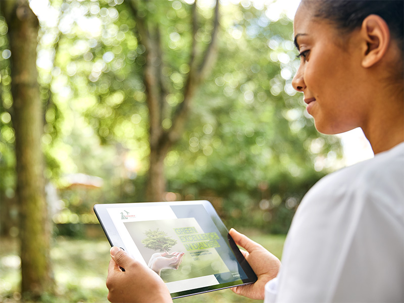 A dialysis nurse standing in a park while ​ viewing the Green Excellence in Dialysis booklet on an iPad.​