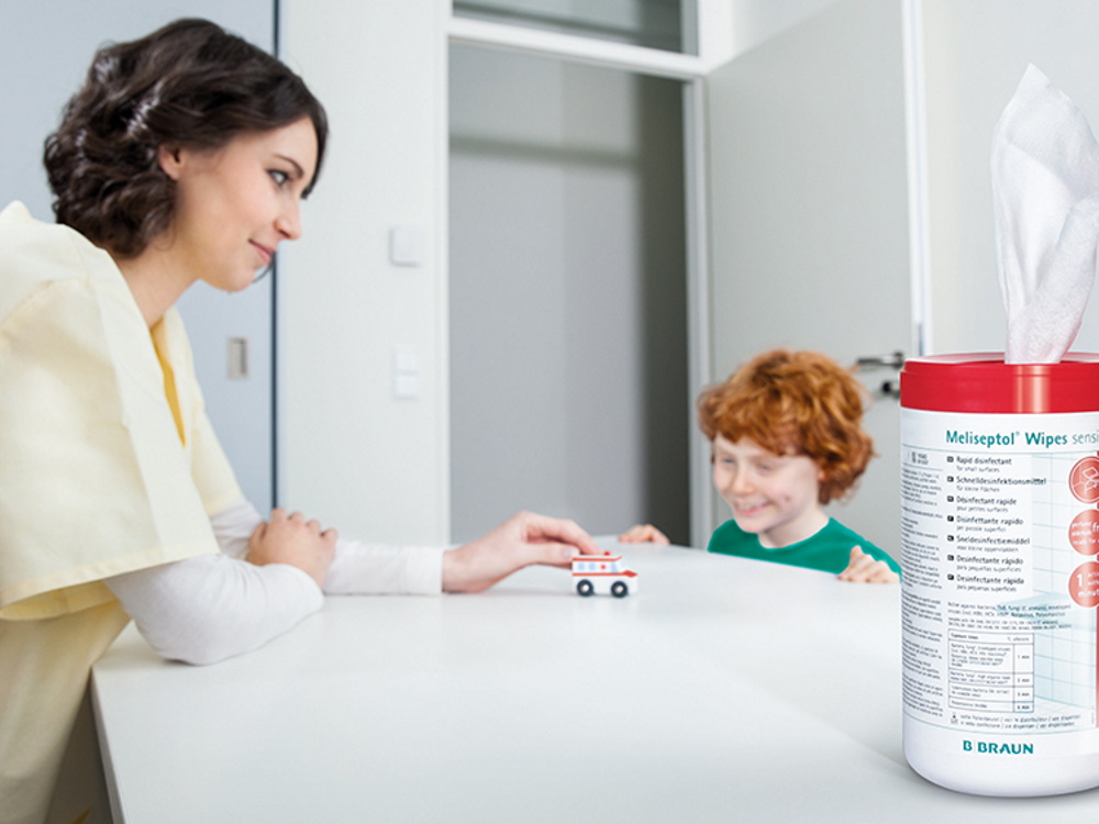 Female nurse standing at a table, opposite a small smiling boy. On the table is a small model ambulance being pushed by the woman, watched by the boy.  In the foreground a pack of Meliseptol Wipes.