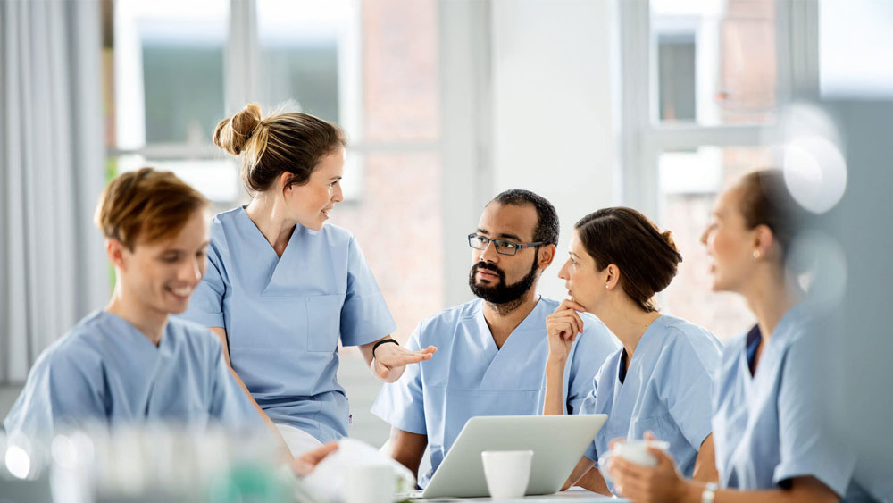 Health care team in light blue scrubs sitting around a table in a bright hospital setting, discussing patient care with a laptop and coffee cups on the table