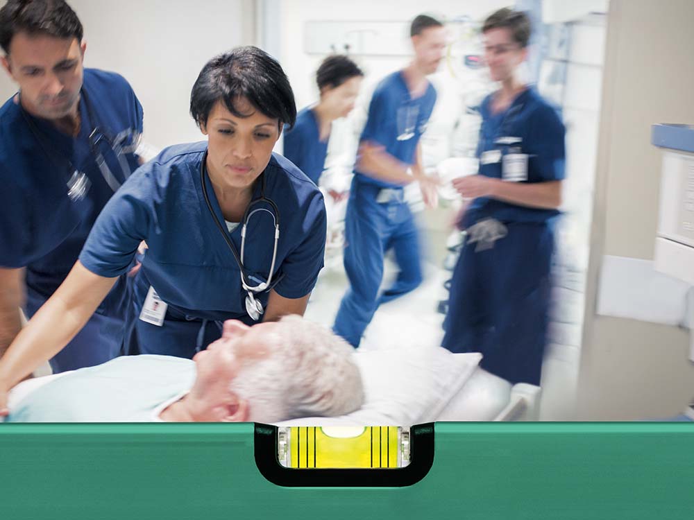 A medical team in clinical workwear is accompanying a person on a hospital bed with acute sepsis through a clinical corridor. In the foreground, a green measuring tool with an integrated spirit level is visible. In the background, medical equipment and monitoring devices are positioned along the wall. ​