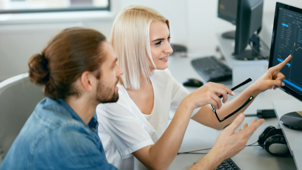 A woman is sitting in front of a computer, pointing at the screen and showing to a man who is sitting next to her.