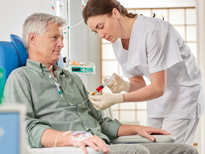 An elderly man is receiving medications through a syringe and tubes from a nurse in a hospital room
