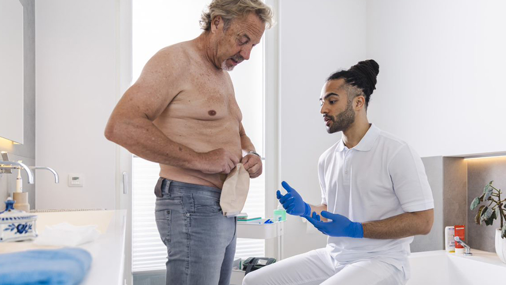 Man with bare upper body in private bathroom manipulates stoma bag on his stomach. Male nurse sits on the edge of the bathtub and gives instructions.