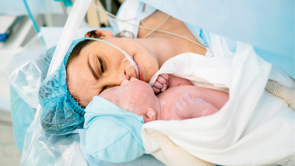 A newborn on a white medical sheet beside a woman in a hospital, likely just after delivery. The mother wears a blue surgical cap and drape, with monitoring wires attached, highlighting a sterile setting and the immediate post-birth moment. 