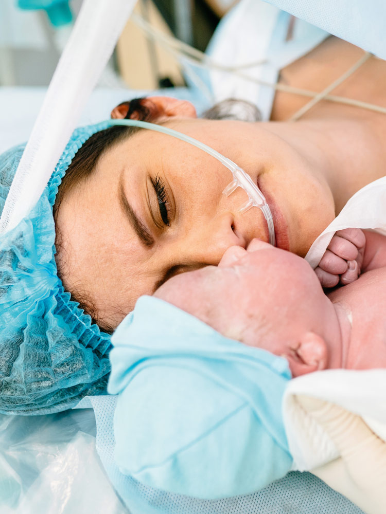 A mother lies in the operating room after a  caesarean section and looks at her newborn baby,  which a nurse is showing her. She looks exhausted,  but also relieved. She is wearing a surgical cap over  her dark hair, the newborn is wrapped in a white  cloth and is wearing a light blue cap.