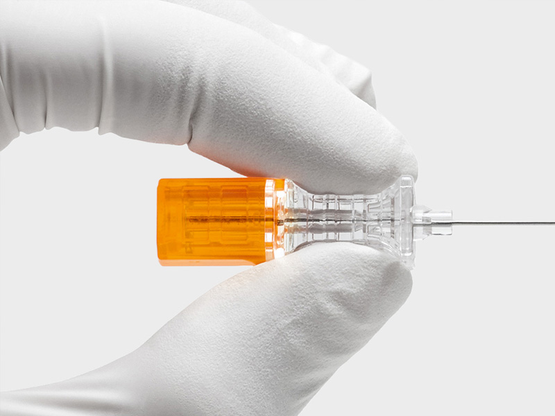 Close-up of a gloved hand holding a Pencan spinal needle with an orange hub and transparent body, used for medical procedures