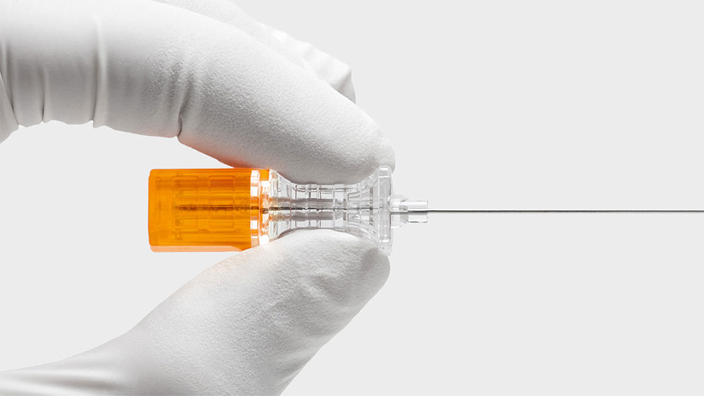 Close-up of a gloved hand holding a Pencan spinal needle with an orange hub and transparent body, used for medical procedures.