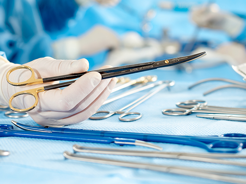 Various surgical instruments on a table in the foreground a hand with a black needle holder