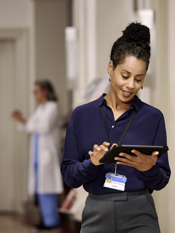 A woman in the foreground, dressed in a dark blue shirt and wearing an identification badge on a lanyard, is standing in a hallway, holding and looking at a tablet. In the background, another person is visible, dressed in medical attire.