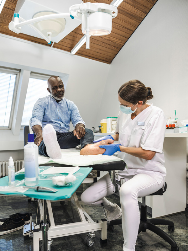 A healthcare professional wearing gloves treats a patient’s foot on an examination table in a clinical room, with medical supplies on a nearby tray and an overhead examination light above.