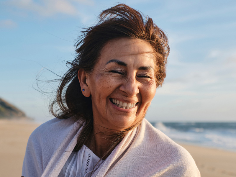Person standing on a sandy beach with ocean waves in the background, wearing a light shawl and a white top, with wind blowing through the hair.