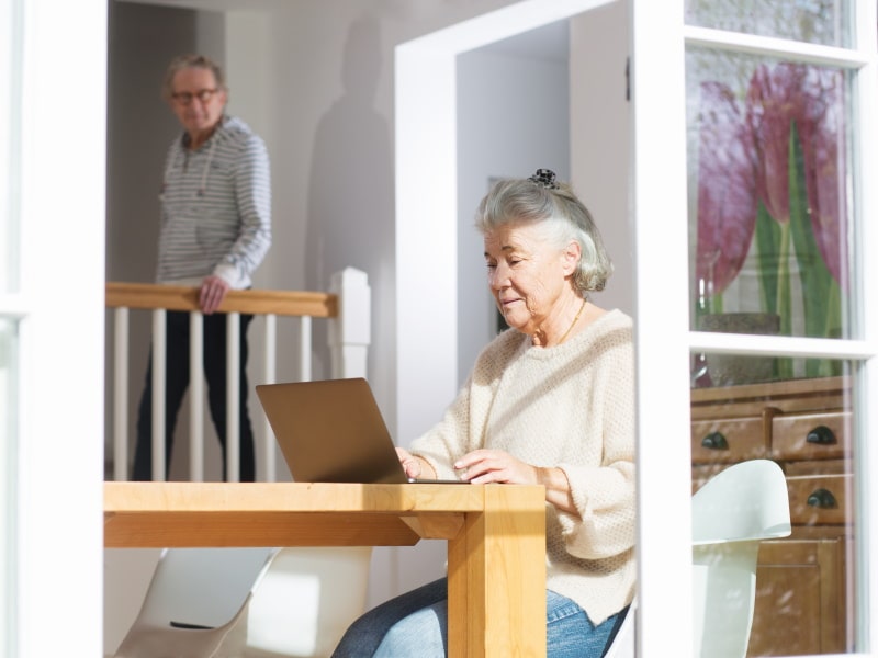 An elderly woman sits at a table, working on a laptop as sunlight streams in around her.