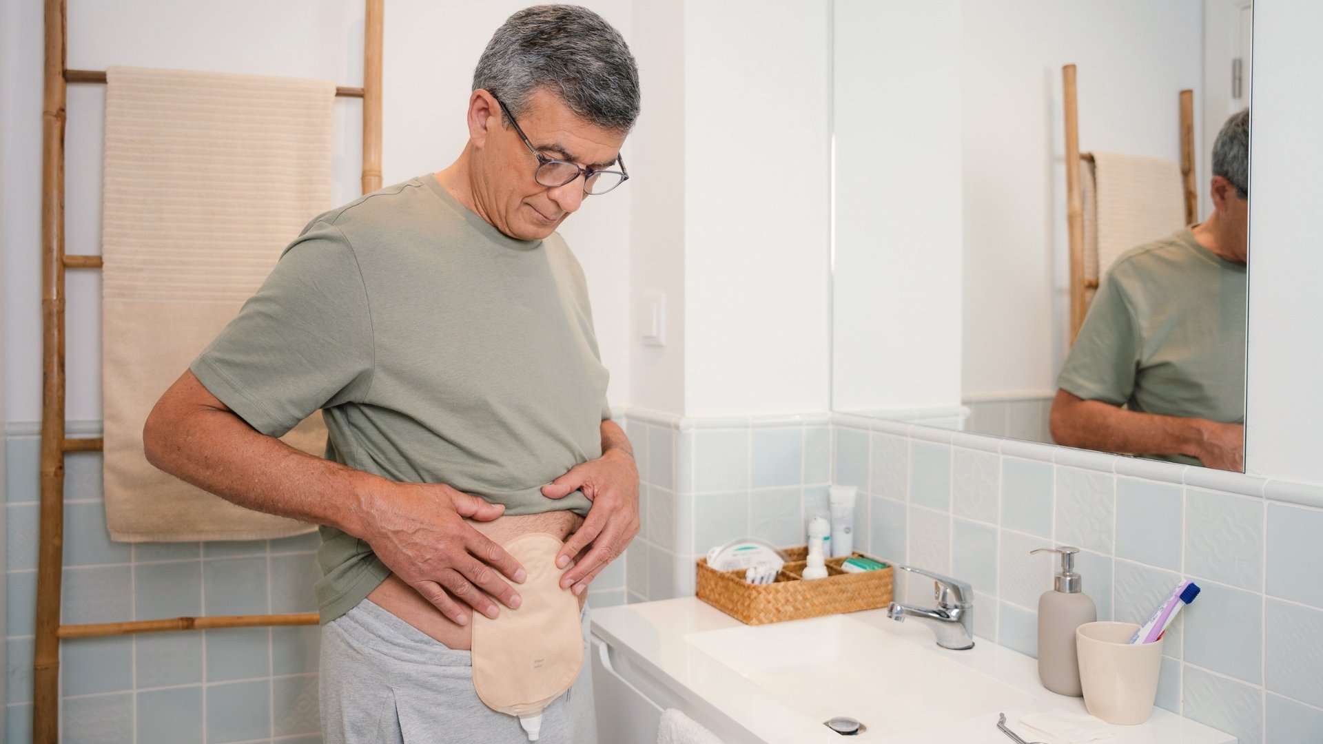 A man wearing glasses and a green t-shirt looking down at his urostomy bag attached to his abdomen while standing in a bathroom. He has with his reflection is visible in a mirror in his side.