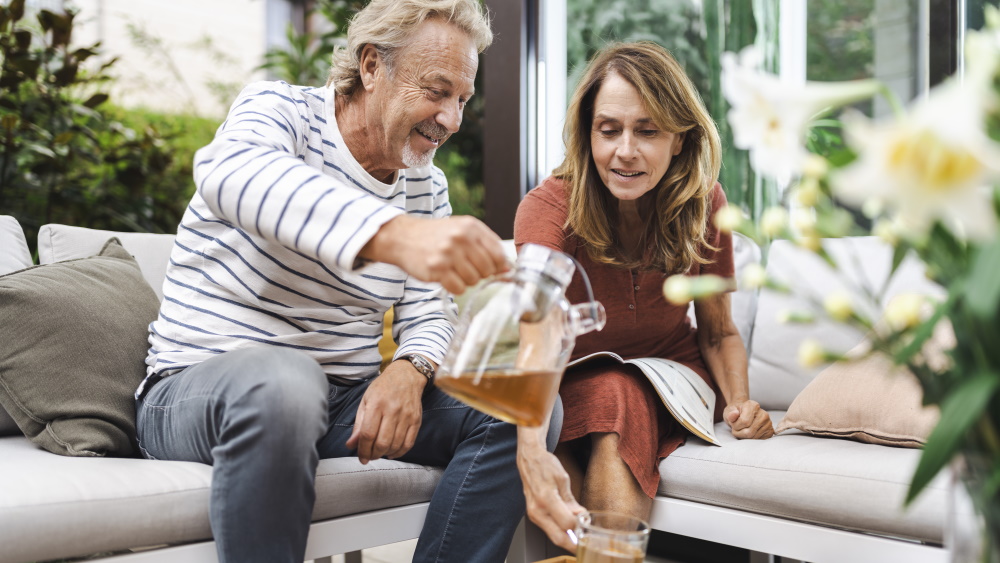A middle-aged man and a middle-aged woman sit together on the sofa and the man pours the woman a cup of tea