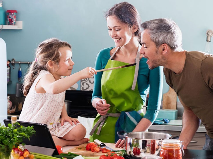 A man, a woman, and a little girl are cooking together. The little girl gives the man a spoon to try the dish.
