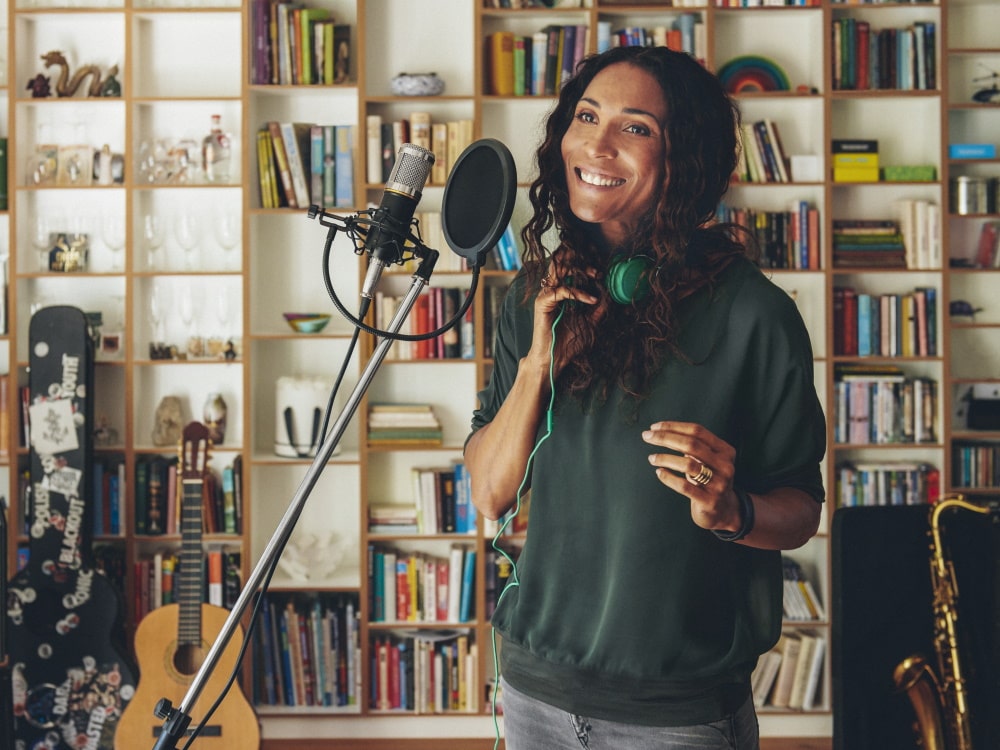 A woman is standing in front of a microphone in a home recording studio, with musical instruments in the background