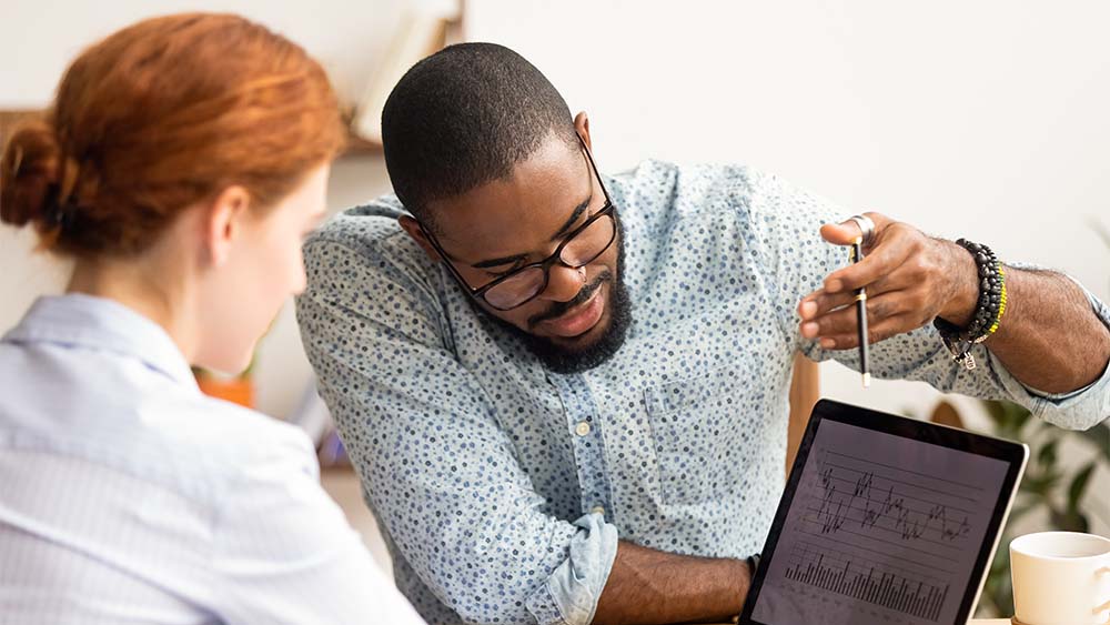 Two individuals discussing business graphs and charts on a tablet in an office room. 