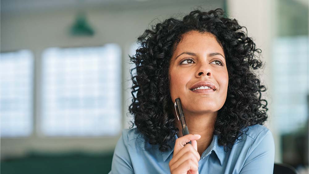 A person with thoughtful expression, holding a pen close to their chin, sitting in an office room. 