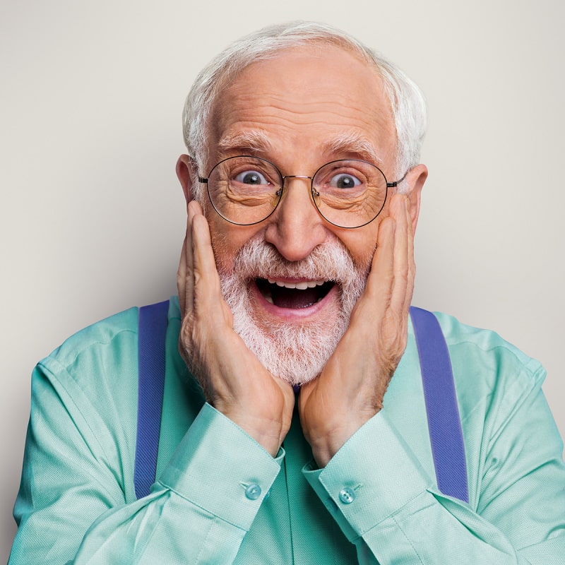 Elderly man with glasses and beard who looks surprised and puts both hands to his face