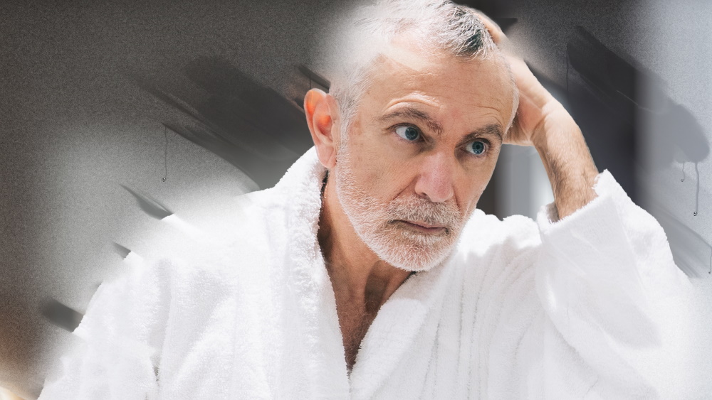 elderly man in bathrobe looks thoughtfully into fogged bathroom mirror