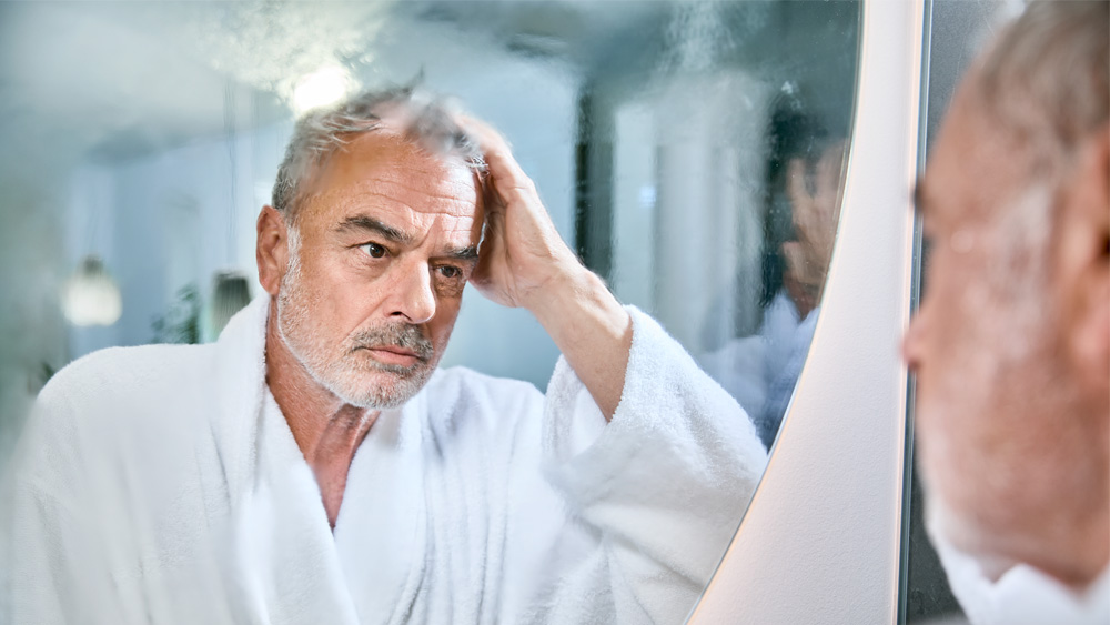 elderly man in bathrobe looks thoughtfully into fogged bathroom mirror